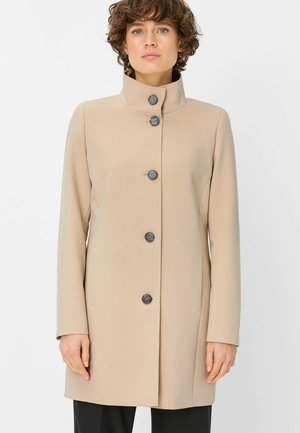 Woman with short curly hair wearing a beige, buttoned-up coat with a high collar, standing against a plain light background.
