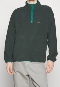 Mørkegrøn fleecepullover med høj krave, frontknapper, to store lommer og kontrastgrønt trim langs bærestykkerne og den nederste kant.