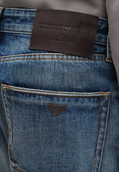 Jeans de mezclilla azul desgastados con costuras naranjas; presentan una etiqueta de cuero marrón oscuro con el logotipo y la inscripción "Emporio Armani" en el bolsillo trasero.