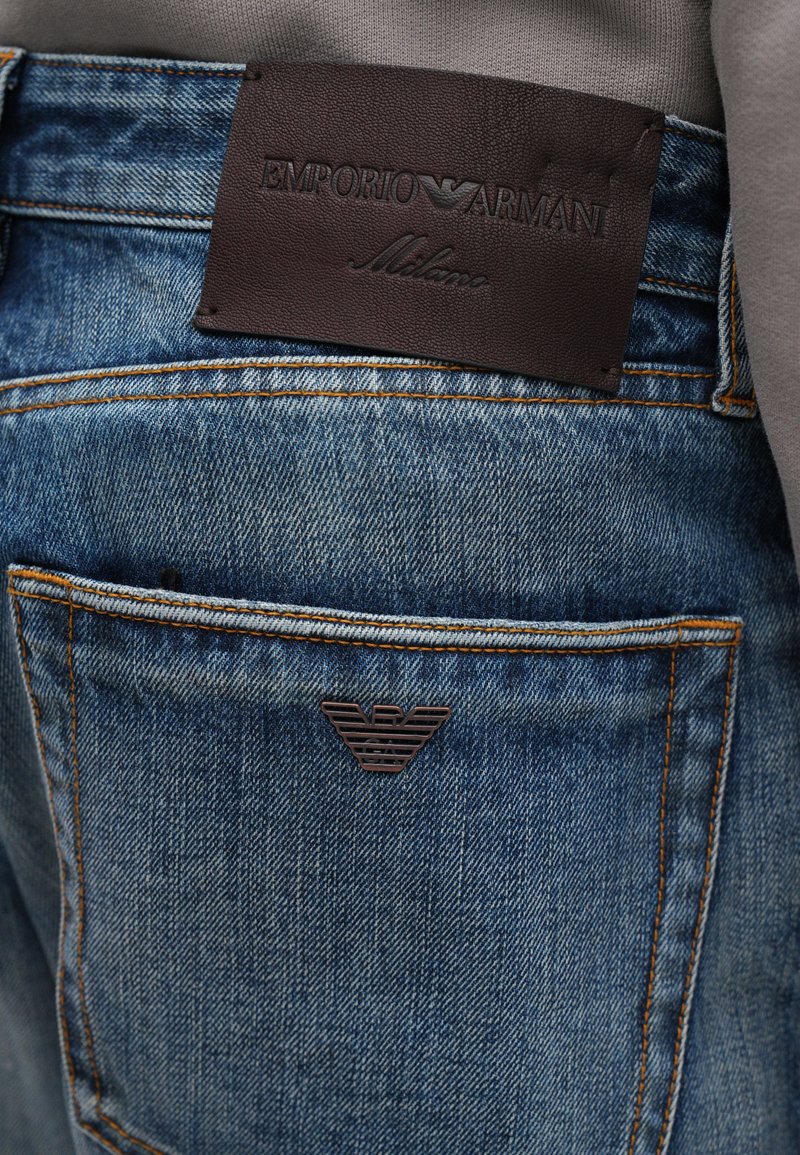 Jeans en denim bleu délavé avec des coutures orange ; présente une étiquette en cuir marron foncé embossée avec "Emporio Armani" et le logo sur la poche arrière.