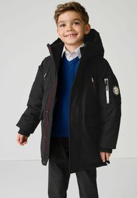 Manteau d'hiver noir avec une capuche, accents rouges, poches zippées et un patch de logo sur la manche. Porté sur un pull bleu et une chemise blanche.