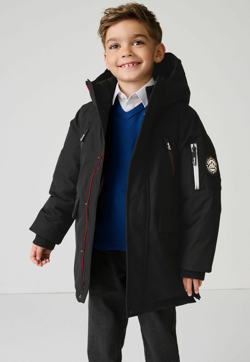 Manteau d'hiver noir avec une capuche, accents rouges, poches zippées et un patch de logo sur la manche. Porté sur un pull bleu et une chemise blanche.