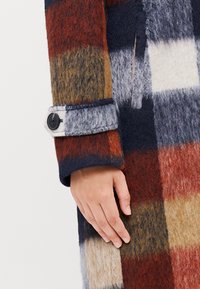 Cappotto di lana con un motivo a quadri multicolore in blu navy, rosso, marrone e crema; presenta una consistenza morbida e un bottone nero sul polsino.