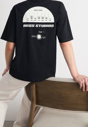 Person trägt ein schwarzes T-Shirt mit der Aufschrift „Boss Studios“ und einer Grafik des Audio-Eingangspegels, lehnt sich an die Rückenlehne eines Holz- und Stoffstuhls.