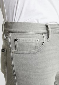 Světle šedé džíny z denimu s pětikusovým designem, viditelným prošíváním a kovovými nýty. Detailní pohled zaměřený na pas a boční kapsu.