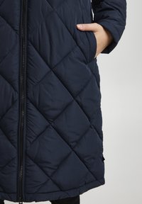 Navy quilted long coat with diamond stitching, featuring a front zipper and side pockets. Made of smooth, lightweight material.