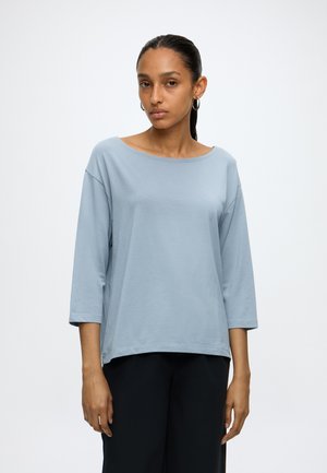 Frau mit glatten, dunklen Haaren im Pferdeschwanz trägt ein lockeres, hellblaues Langarmshirt und schwarze Hose und steht vor einem schlichten Hintergrund.