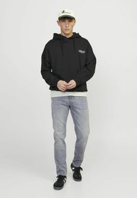 Sweat à capuche noir avec cordons, poche avant et logo. Porté avec un jean gris clair et des baskets noires, complété par une casquette beige.