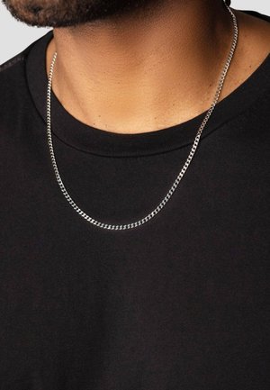 Sølv kædetrøje med curb-links, hvilende på en bar hals, over en sort t-shirt med rund halsudskæring.