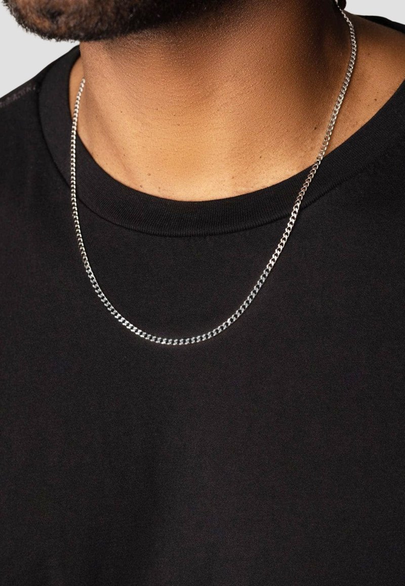 Sølv kædetrøje med curb-links, hvilende på en bar hals, over en sort t-shirt med rund halsudskæring.