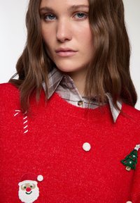 Czerwony sweter z dzianiny o okrągłym dekolcie, z haftowanym mikołajem, choinką i akcentami w postaci cukrowych lasek. Warstwowy na koszuli w kratę z kołnierzykiem.