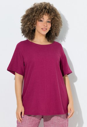 Femme aux cheveux bouclés portant un haut ample à manches courtes magenta et un pantalon rayé rose, debout sur un fond blanc.