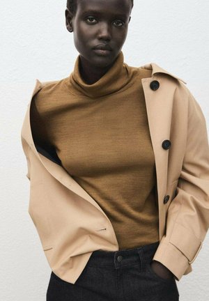 Persona de piel oscura con un suéter de cuello alto marrón claro, abrigo beige con botones negros y pantalones negros, de pie contra una pared blanca lisa.