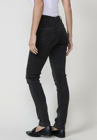 Lieknos juodos džinsinės kelnės su lengvu blizgesiu, vidutinio aukščio juosmuo ir dvi galinės kišenės. Paprasta siuvimo forma ir kontrastingos metalinės detalės.