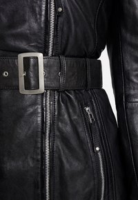 Svart läderjacka med en slät yta, silverfärgade dragkedjedetaljer, ett brett bälte med en rektangulär spänne och sidofickor med dragkedja.