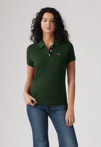 Donkergroene poloshirt van katoen met korte mouwen, een platte kraag en een knoopsluiting met drie knopen. Rode logo op de borst. Draagt met blauwe spijkerbroek.