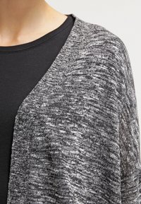 Persona vestida con una camiseta negra de cuello redondo debajo de un cárdigan de punto jaspeado en gris y blanco, mostrada desde el pecho hasta el hombro.