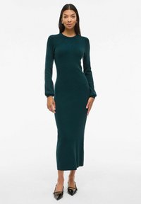 Vestido de punto de manga larga en azul oscuro con una silueta ajustada, escote redondo y textura suave. Combinado con zapatos negros de punta.