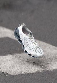 White athletic shoe with textured upper, grey accents, and a distinctive cushioned sole featuring circular cutouts. Floating above a surface.