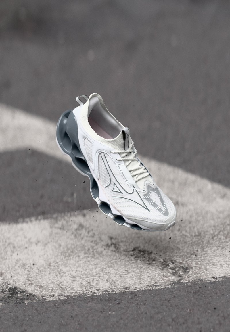 White athletic shoe with textured upper, grey accents, and a distinctive cushioned sole featuring circular cutouts. Floating above a surface.