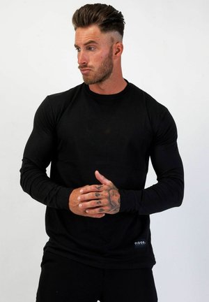 Muskelspänd man med stylat hår och handtatueringar, iklädd en svart tröja med långa ärmar, tittar åt sidan med sammanlänkade händer.