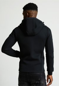 Crna majica s kapuljačom s prilagodljivim dizajnom, teksturiranim materijalom i kapuljačom. Značajke uključuju diskretne detalje šavova i rebraste manžete.