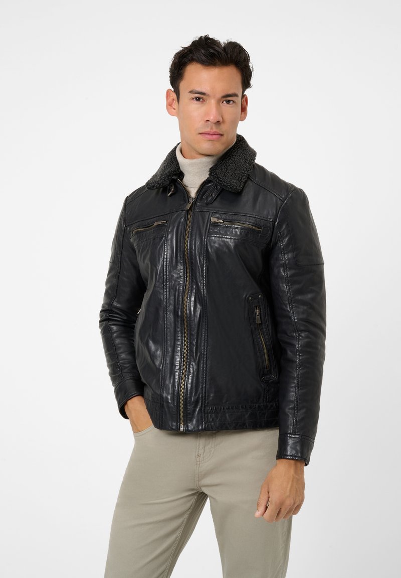 Svart läderjacka med en krage i syntetpäls, dragkedjefickor och en figurnära design. Modellen har på sig en ljusgrå polo och beige byxor.