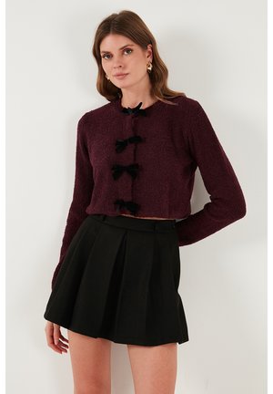 Cardigan court bordeaux avec des manches longues, en tissu texturé, et trois accents de nœud noir ; associé à une jupe noire plissée taille haute.