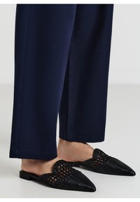 Mules tissés noirs avec des orteils pointus et une finition texturée, associés à un pantalon large bleu marine, mettant en valeur un design épuré et moderne.