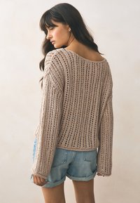Strikket sweater i lys beige med et løst, åbent vævemønster. Har en bred halsudskæring og lange ærmer, kombineret med denimshorts.