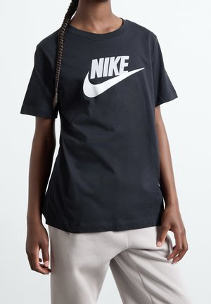 Osoba o ciemnych zaplecionych włosach, ubrana w czarną koszulkę Nike z białym logo i jasne spodnie, stojąca na tle jednolitego tła.