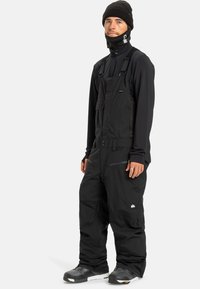 Man in zwarte sneeuw-broek, zwart langmouwtop, zwarte beanie, nekkraag en zwarte sneeuwlaarzen staat tegen een witte achtergrond.