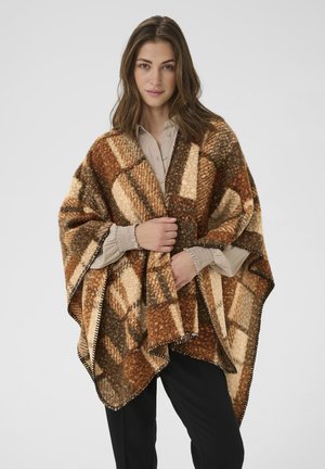 Femme portant un blouse beige et un pantalon noir, drapée dans un châle à carreaux texturé marron, beige et crème, se tenant devant un fond uni.