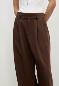 Bruna skräddarsydda byxor med hög midja, veckad design och sidofickor, tillverkade i ett slätt tyg med subtil glans.