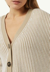 Beiger Cardigan mit V-Ausschnitt, der weiße strukturierte Streifen und große braune Knöpfe vorne aufweist. Weiches Strickmaterial.
