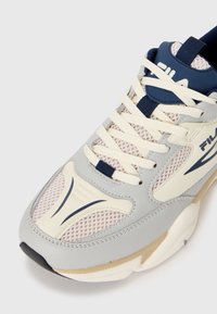 Närmre bild av en FILA-sneaker med beige mesh, grå och krämfärgade överlägg, krämfärgade skosnören och marinblå accenter på tungan och logotypen.