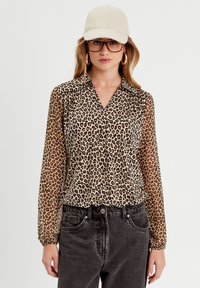 Blusa de leopardo con mangas largas, diseño con cuello, tela transparente y detalles en negro. Combinada con jeans de mezclilla oscuros.