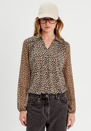 Blouse imprimé léopard avec des manches longues, design col, tissu transparent et accents noirs. Associée à un jeans en denim foncé.