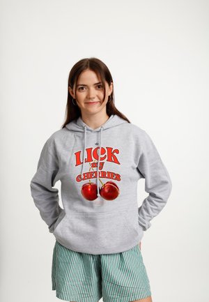 Młoda kobieta o brązowych włosach, ubrana w szarą bluzę z kapturem z czerwonymi wiśniami i napisem, w zielono-białe paskowane szorty, stoi z rękami za plecami.