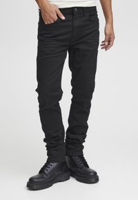 Črne ozke jeans hlače s gladko teksturo, z gumbi spredaj in stranskimi žepi, kombinirane s črnimi škornji z vezalkami.