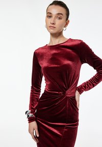 Bordowa aksamitna sukienka z długimi rękawami, z wykręconym detalem w talii i gładką fakturą. Srebrna bransoletka na nadgarstku.