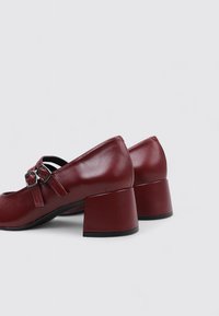 Zapatos de tacón de cuero burdeos con un tacón cuadrado robusto y acentos de hebilla. Textura suave con un diseño de punta redonda.
