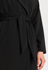 Sort, langærmet omslagsbadekåbe med bindebælte, der har en glat tekstur og en afslappet pasform. Synlig lomme og draperede stofdetaljer.