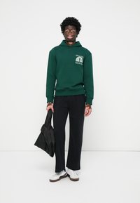 Grøn hoodie med frontlomme og hvidt logo, parret med sorte bukser og en sort jakke. Hvide sneakers med brune såler.