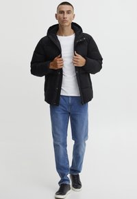 Doudoune noire avec capuche, dotée d'une fermeture éclair et de boutons-pression. Portée sur une chemise blanche avec un jean bleu et des baskets noires.