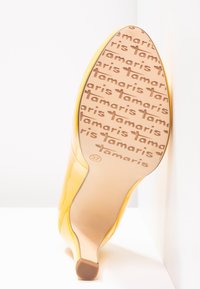 Chaussure à talon high heel en cuir verni jaune, inclinée pour montrer la semelle avec le logo "Tamaris" répété et la taille 37.