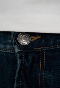 Prim-plan al unui nasture metalic care fixează o pereche de blugi din denim albastru închis sub tivul unui articol vestimentar alb cu detalii vizibile de cusătură.