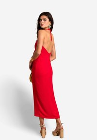 Rotes Halfterkleid mit offenem Rücken, figurbetontem Design und knielangem Saum. Getragen mit klobigen Plateausandalen und auffälligen Ohrringen.