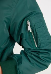 Zelena bomber jakna s glatkom teksturom. Ima zippered džep na rukavu i rebraste manžete. Lagani i uski dizajn.