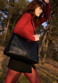 Žena v červenom tričku a legínach kráča vonku, nesúc veľkú čiernu koženú kabelku cez rameno v lesnatej oblasti počas denného svetla.